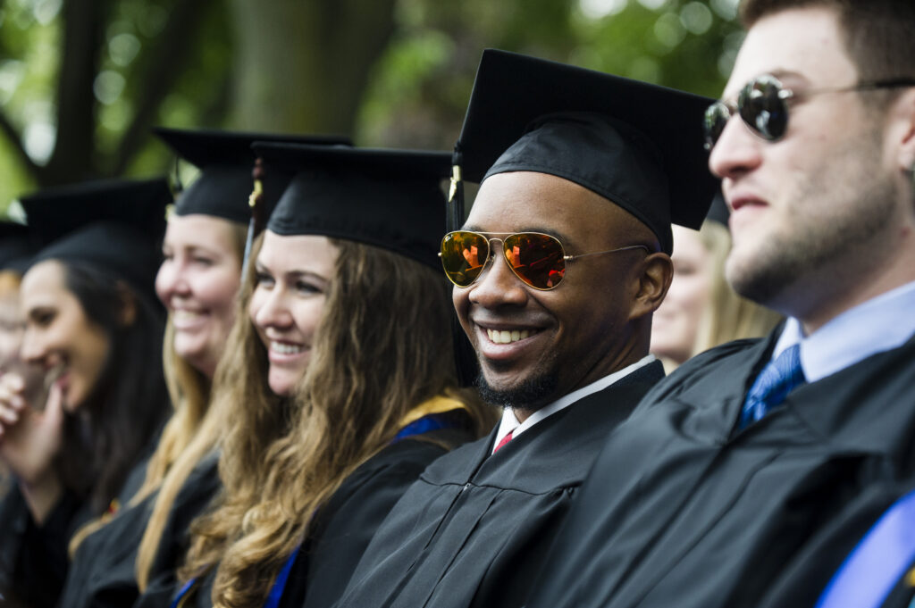 Graduating students wearing caps and gowns sit in a row, smiling.
