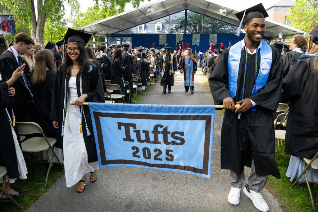 Graduates walk during Commencement while holding a Tufts 2025 banner.