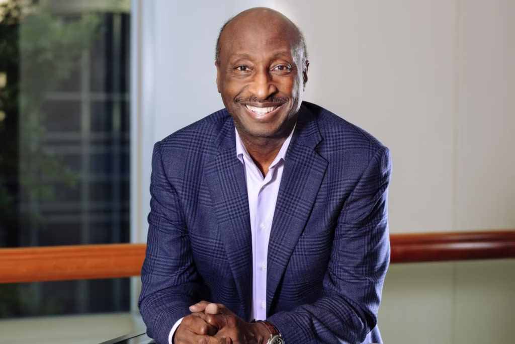 Ken Frazier, wearing a blue blazer, leans on a glass table looking forward
